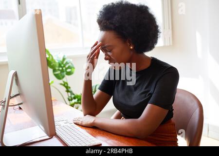 Tired and upset young developer sits with head in hands at desk, Black woman Stock Photo