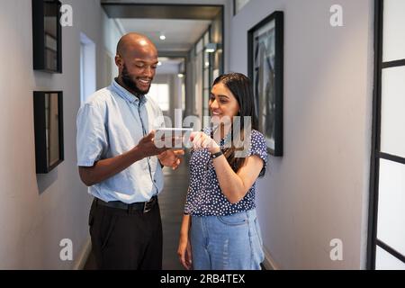 Two colleagues stand in passage hallway, discussing work with digital tablet Stock Photo