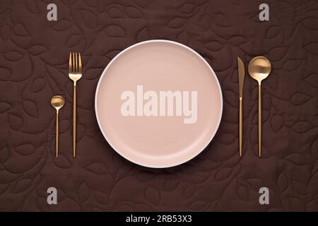 Festive table setting on dark tablecloth. Empty plate and gold cutlery on a brown background. Top view. Dining table in luxury restaurant. Card or men Stock Photo