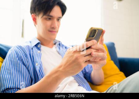 Portrait of young man using mobile phone and sitting on couch in living room, Happy smart man relaxing and using smartphone on sofa at home on weekend Stock Photo