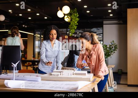 Diverse female architects in discussion using model and blueprints in casual office meeting. Casual office, teamwork, business, lifestyle, creative wo Stock Photo