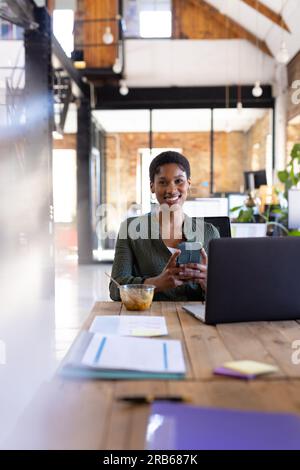 Portrait of happy african american casual businesswoman using smartphone and having lunch in office. Business, food, technology and work, unaltered. Stock Photo