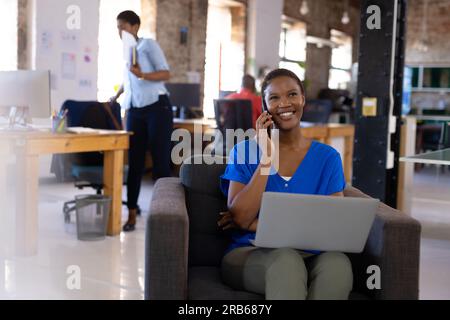 Happy african american colleagues using computer and talking on smartphone in creative office. Business, technology and teamwork, unaltered. Stock Photo