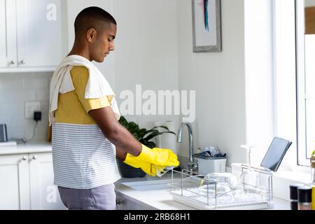 Biracial man washing up in kitchen sink and using tablet with copy space on screen. Healthy lifestyle, chores, cleaning, communication and domestic li Stock Photo