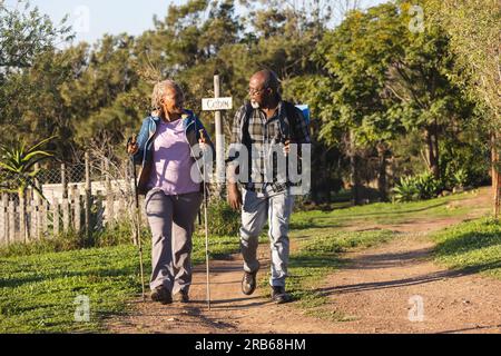 Happy senior african american couple with backpacks walking in sunny nature. Senior lifestyle, relaxing, wellbeing in nature and retirement, unaltered Stock Photo