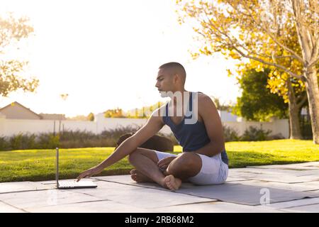 Focused biracial man practicing yoga sitting in sunny garden using laptop. Summer, wellbeing, communication, fitness and healthy lifestyle, unaltered. Stock Photo