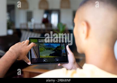 Biracial man sitting in living room using tablet with home security camera views on screen. Technology, communication, security, lifestyle and domesti Stock Photo