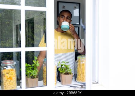 Thoughtful biracial man drinking coffee standing in kitchen, seen through window, copy space. Free time, wellbeing, lifestyle and domestic life, unalt Stock Photo