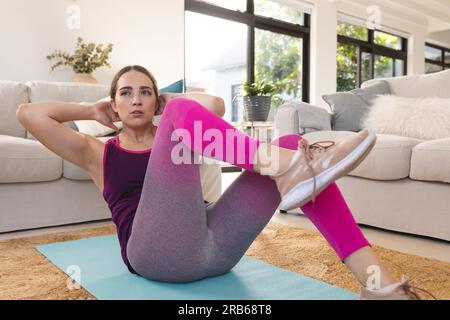 Focused caucasian woman exercising, doing sit ups on mat at home. Domestic life, fitness, activity and lifestyle, unaltered. Stock Photo