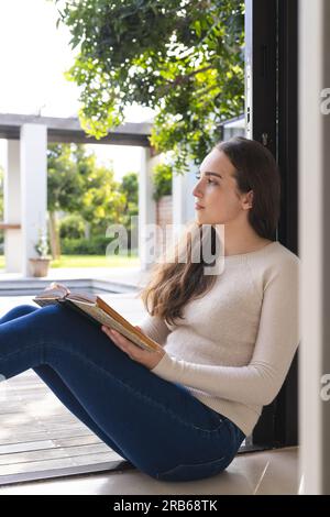 Thoughtful caucasian woman sitting at wall and holding book at home, copy space. Domestic life, relaxation and lifestyle, unaltered. Stock Photo