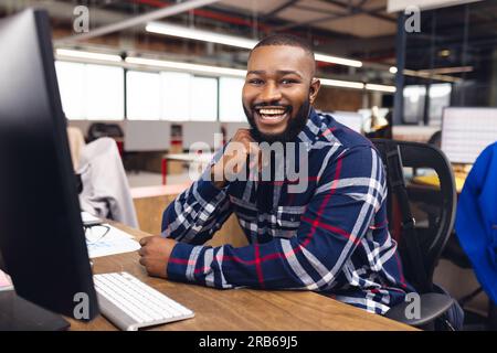 Portrait of happy african american businessman using computer and smiling at office. Casual business, confidence, communication and work, unaltered. Stock Photo