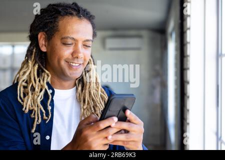 Happy biracial casual businessman with dreadlocks using smartphone by window in sunny office. Work, business, communication and casual office, unalter Stock Photo