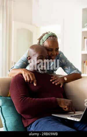 Happy african american senior couple using laptop and embracing in sunny living room. Senior lifestyle, retirement, togetherness, communication and do Stock Photo