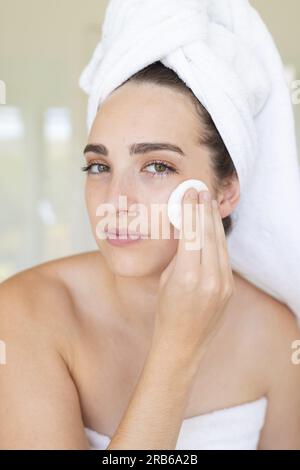 Portrait of caucasian woman with towel on head using pad on face in bathroom. Domestic life, selfcare and lifestyle, unaltered. Stock Photo