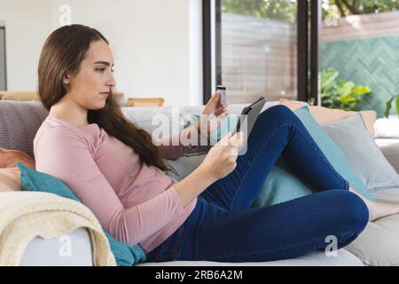Focused caucasian woman on couch using tablet and holding credit card at home. Domestic life, shopping, technology and lifestyle, unaltered. Stock Photo