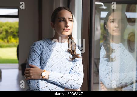 Thoughtful caucasian woman looking through window with arms crossed at home. Domestic life and lifestyle, unaltered. Stock Photo