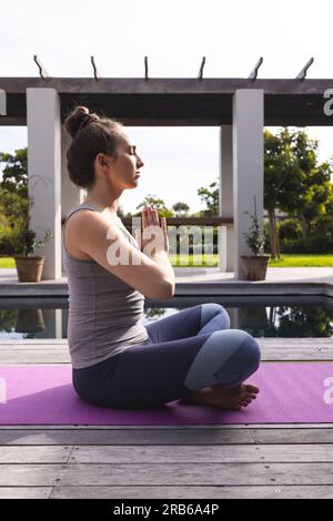 Focused caucasian woman on mat practicing yoga and meditating in garden. Domestic life, fitness, activity and lifestyle, unaltered. Stock Photo