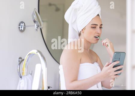 Happy caucasian woman with towel on head brushing teeth and using smartphone in bathroom. Domestic life, technology, selfcare and lifestyle, unaltered Stock Photo