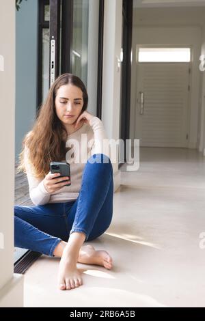 Serious caucasian woman sitting at wall and using smartphone at home. Domestic life, technology and lifestyle, unaltered. Stock Photo