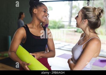 Happy diverse women holding mat and talking in yoga class. Yoga, exercise, fitness and healthy lifestyle, unaltered. Stock Photo