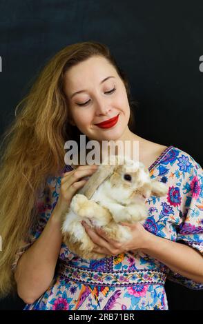 Beautiful russian woman with long blond hair holding a rabbit pet in her hands against black wall Stock Photo