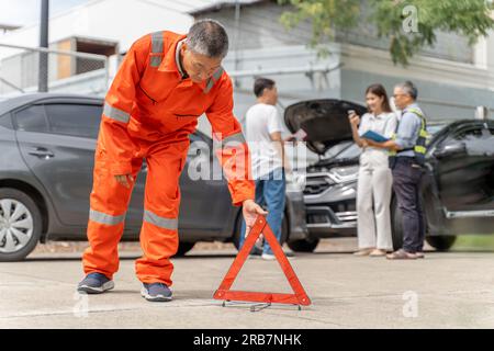 A car insurance agent in orange jumpsuit placing a warning triangle down on the road near the car accident while another insurance agent talking to th Stock Photo