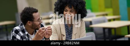 Two young happy mixed race colleagues sitting at the desk in coworking space, using laptop and discussing new project, guy explaining something to his Stock Photo