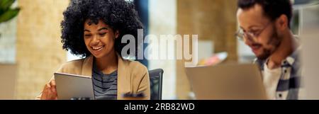 Two young happy multiracial business people sitting at the desk, using modern technologies while working together in coworking space Stock Photo
