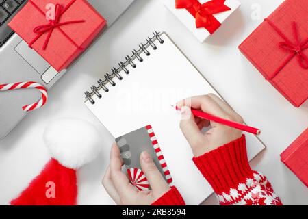 Christmas online shopping flat lay. Girl writing shopping list. Female Hands with Credit card and Pencil. Laptop and christmas presents on white table Stock Photo