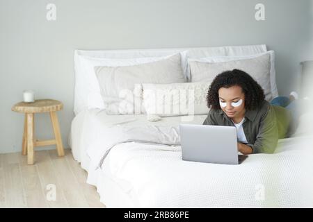 African-American teen with under-eye patches works on laptop in bed. Biracial lady shops online during morning skincare. Stock Photo