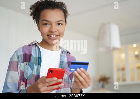 Happy biracial teen pays with credit card, smartphone. Using online banking, e-bank apps. Secure purchase, money transfer at home. Stock Photo