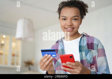 Happy biracial teen pays with credit card, smartphone. Using online banking, e-bank apps. Secure purchase, money transfer at home. Stock Photo