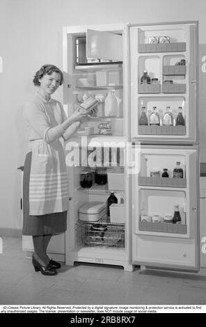 In the kitchen 1960s. A woman in her  kitchen with the door open to the the refrigerator. You can see the food products of various kinds that are neatly displayed on the shelves. Among other things, the classic Tetran milk package is visible. Invented by Ruben Rausing and which became the start of the Tetra Pak packaging empire. Sweden 1960. Conard ref 4188 Stock Photo