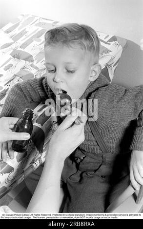 A boy is sick and won't take his medicine. His mother holds out a spoon and waits for him to gape and take it, and he does. He opens his mouth and takes it even though it for sure does not tase so good. Sweden 1956. Conard ref 3157. Stock Photo