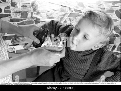 A boy is sick and won't take his medicine. His mother holds out a spoon and waits for him to gape and take it, but he keeps his mouth shut. An everyday life event that today would look the same, the medicine sometimes does not taste so good. Sweden 1956. Conard ref 3157. Stock Photo
