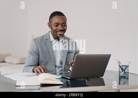 Smiling african american businessman satisfied with his business project, working on laptop, reading email with good news, sitting at office desk. Bla Stock Photo