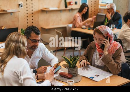 Muslim female employee wearing hijab having phone call and listening to her middle-aged coworkers discussing project details Stock Photo