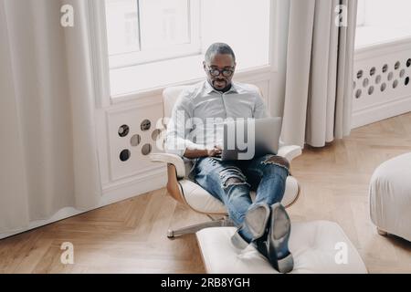 Focused african american businessman works online at laptop, sitting in armchair indoors. Modern black male employee manager in glasses answers email, Stock Photo