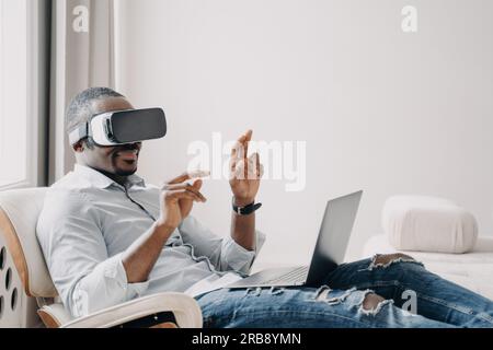 African american male software developer in virtual reality glasses tests new app, working on laptop, sitting in chair. Modern businessman touching ob Stock Photo