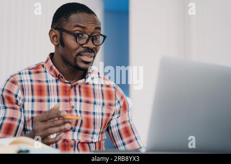 Puzzled african american employee in glasses looking at laptop screen shocked by breakdown of computer or problems in work. Outraged black male is per Stock Photo