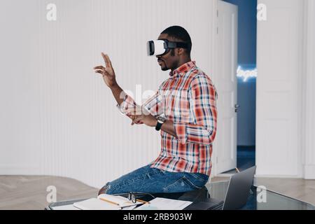 Modern african american guy wearing VR glasses interacts with augmented reality objects, sitting on office desk. Black young man using virtual reality Stock Photo