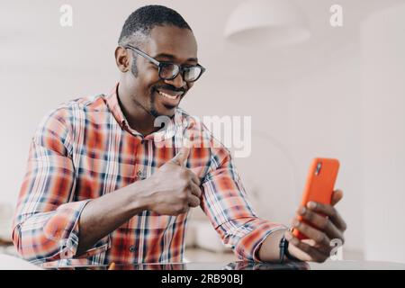Pleased african american man showing thumb up hand gesture, holding smartphone, chatting by video call. Smiling black guy in glasses looking at phone Stock Photo