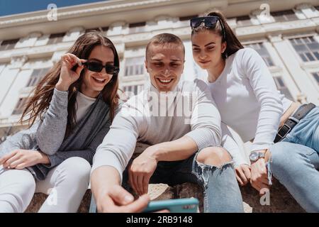Close up of three friends taking a selfie while sitting outside in front of the white building Stock Photo