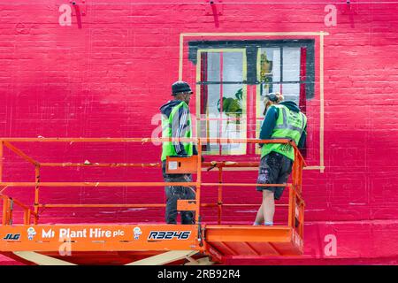 two workers in hi-vifluorescent jackets on high platform by a wall painting art of a window on red painted brick wall on building shoreditch london uk Stock Photo