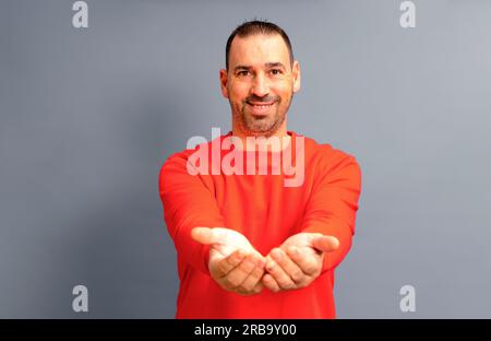 Bearded Hispanic man in his 40s wearing a red jumper offering something imaginary in his clasped hands, isolated on gray studio background. Stock Photo