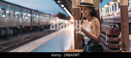 Cheerful young Hispanic Brunette woman sitting on bench on station platform using phone Stock Photo