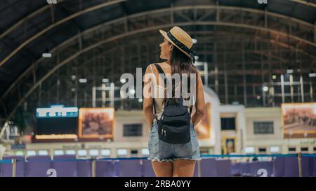 Rear view of young female traveller standing with backpack and hat at train station looking around for sign board to travel towards destination during vacation Stock Photo