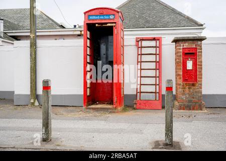 The door of a phone box stands next to the kiosk and a Royal Mail postal box on a coastal road, on 4th July 2023, in Milford-on-Sea, Hampshire, England. Stock Photo