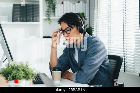 Tired and unhappy customer service operator man with headsets working at call centre, Call center agent with problem on support hotline in modern Stock Photo