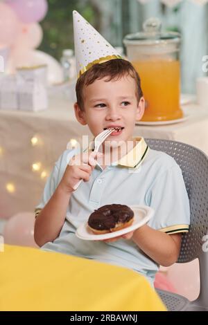 Portrait of cheerful preteen boy eating doughnut with chocolate glaze at birthday party Stock Photo